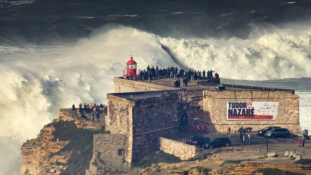 Nazaré: Where the Atlantic Writes Its History | Guide A massive, towering big wave breaking violently in a wall of white water directly in front of the historic stone fort and red lighthouse in Nazaré.