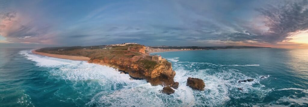 Nazaré: Where the Atlantic Writes Its History | Guide A stunning panoramic aerial drone view of Nazaré, capturing the historic stone fort on the cliffside separating the massive waves of Praia do Norte from the wide golden beach of the town.