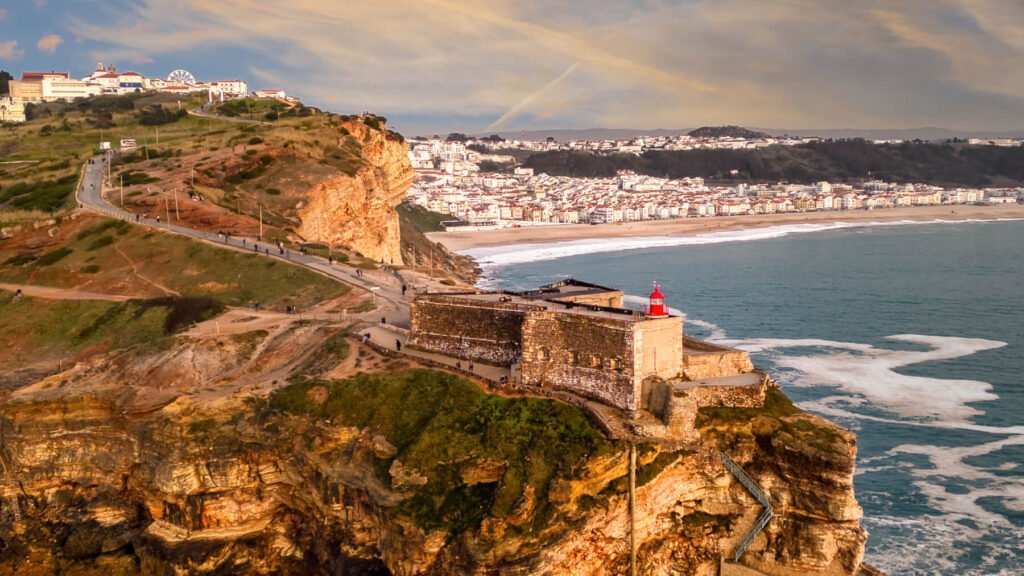 Nazaré: Where the Atlantic Writes Its History | Guide A breathtaking warm sunset aerial drone photograph of the historic stone fort and red lighthouse in Nazaré, with the golden beach and coastal town glowing in the background.