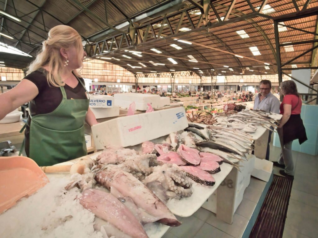 Nazaré: Where the Atlantic Writes Its History | Guide A local Portuguese fishmonger talking to customers behind a market table overflowing with a fresh selection of glistening Atlantic fish inside the Nazaré Municipal Market.
