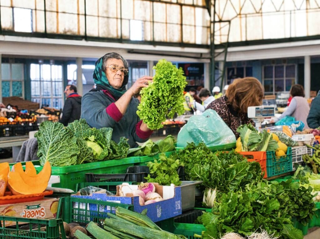 Nazaré: Where the Atlantic Writes Its History | Guide An elderly Portuguese woman with glasses cleaning fresh lettuce at her vibrant produce stall, surrounded by assorted local fruits and vegetables inside the Mercado Municipal da Nazaré.