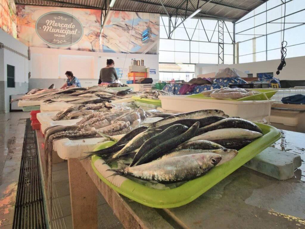 Nazaré: Where the Atlantic Writes Its History | Guide Freshly caught local fish displayed in the foreground, with traditional Portuguese fishmongers cleaning catch at their stalls inside the Mercado Municipal da Nazaré.