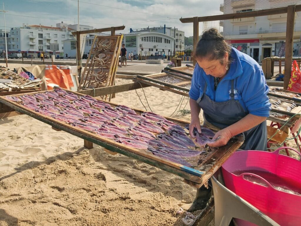 Nazaré: Where the Atlantic Writes Its History | Guide A traditional Portuguese fisherwoman tending to rows of split fish sun-drying on wooden wire racks along the golden beach of Nazaré.