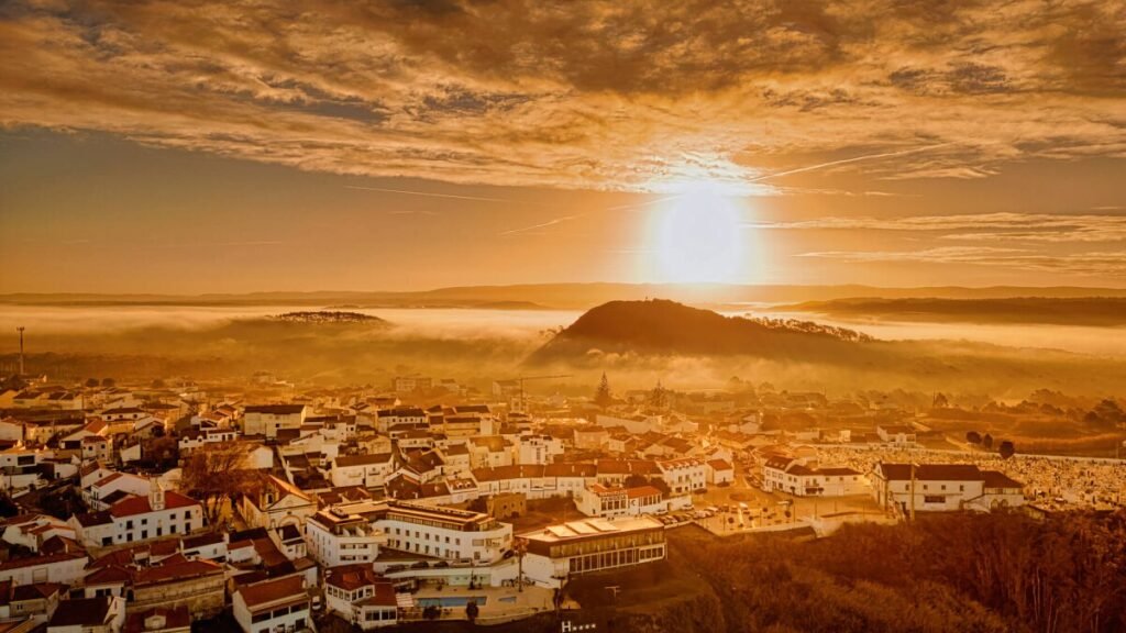 Nazaré: Where the Atlantic Writes Its History | Guide A breathtaking warm sunrise aerial drone photograph looking across the historic hilltop district of Pederneira toward the iconic Monte de São Bartolomeu peak in Nazaré.