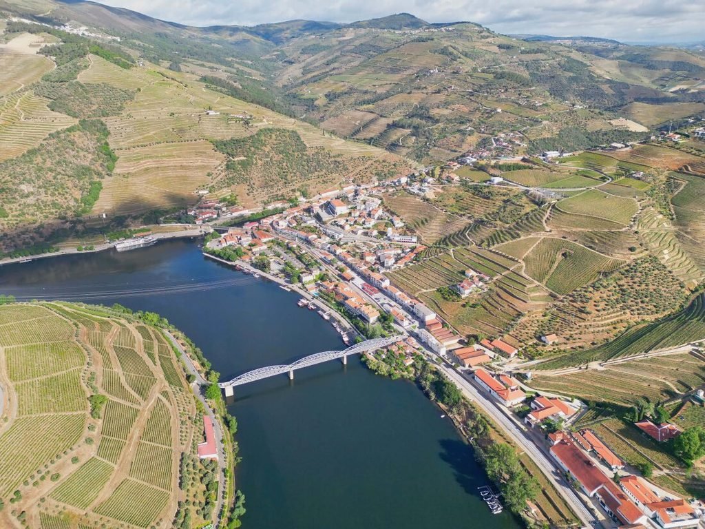 A breathtaking aerial drone photograph looking down at the town of Pinhão, showcasing the historic bridge crossing the Douro River surrounded by steep terraced vineyard mountains under bright daylight.