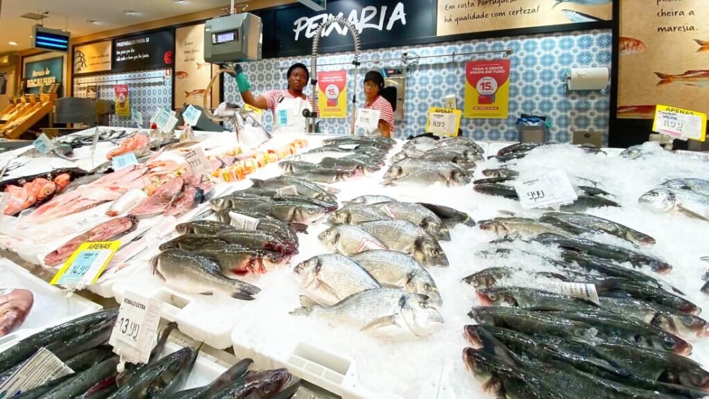 Nazaré: Where the Atlantic Writes Its History | Guide Freshly caught local fish displayed in the foreground, with traditional Portuguese fishmongers cleaning catch at their stalls inside the Mercado Municipal da Nazaré.