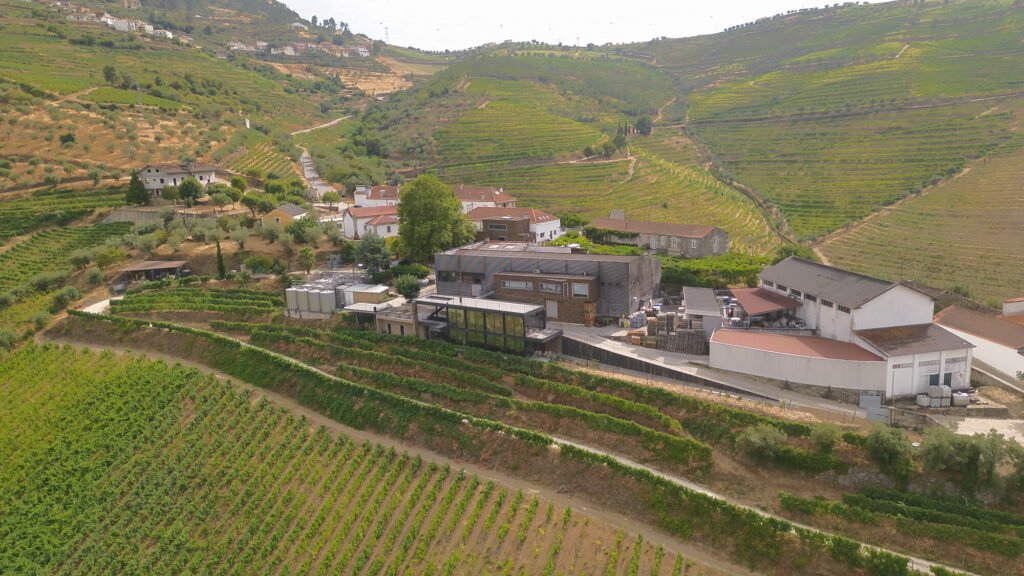 A striking aerial drone photograph looking down at the historic family-run Quinta de Santa Eufémia estate and its modern wine tasting room, surrounded by sprawling terraced vineyards in the Douro Valley.