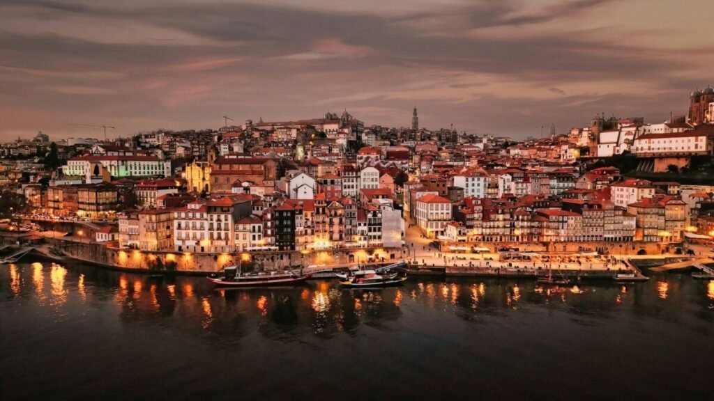 Porto, Portugal: A Slow Travel Guide A stunning aerial drone photograph of the historic Ribeira waterfront district in Porto at twilight, showcasing illuminated city lights reflecting on the calm Douro River.