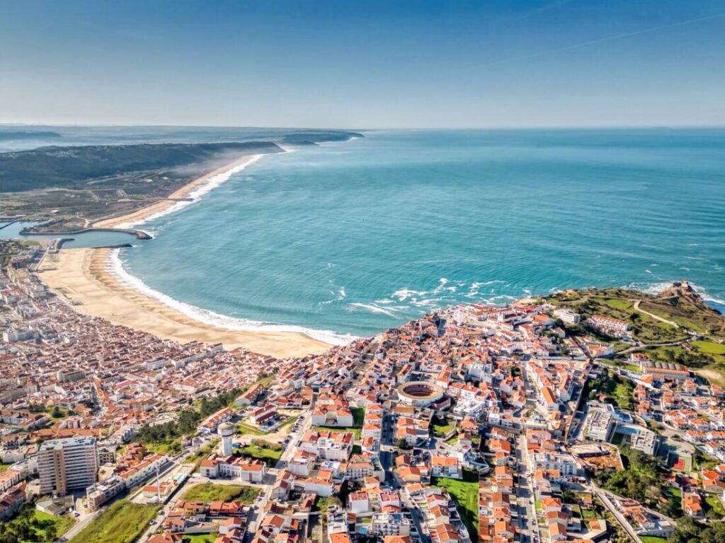 A magnificent aerial drone view captured from behind the Sítio cliffs, overlooking the entire white-walled town and golden crescent beach of Nazaré on a clear sunny day.