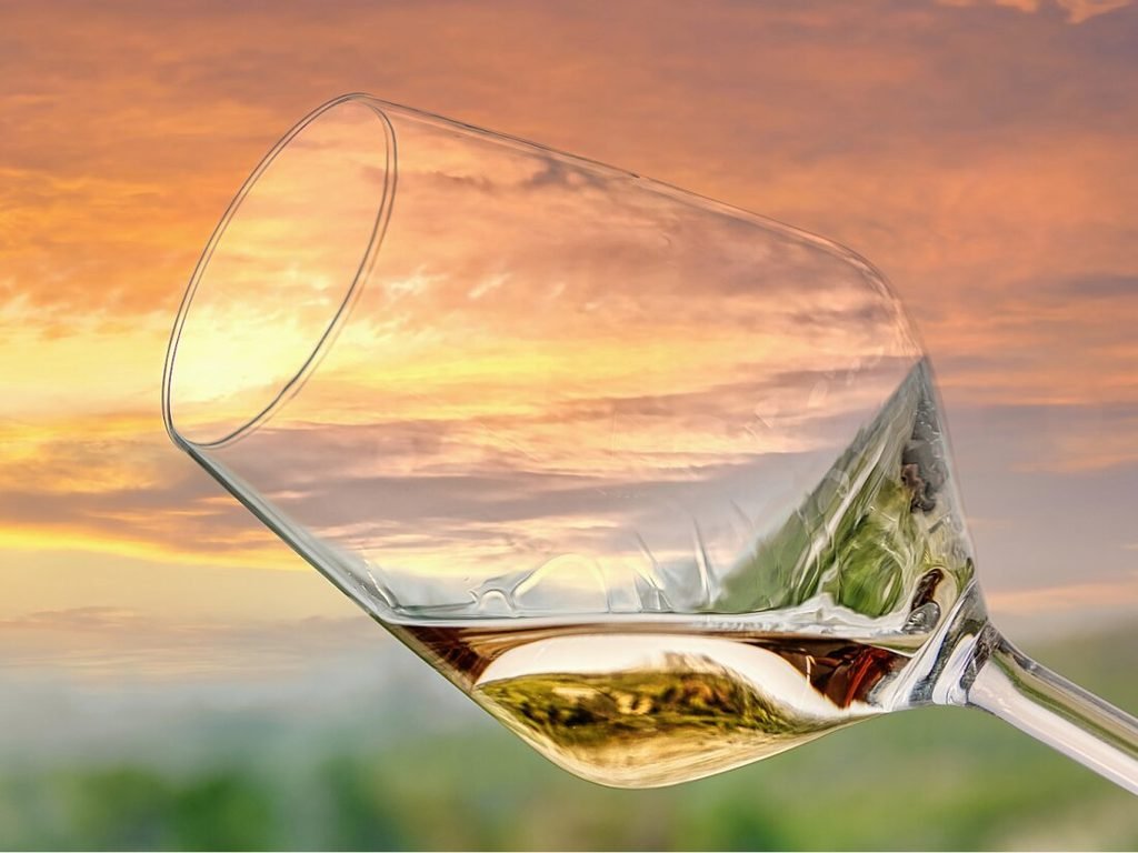 A close-up photograph of a elegant glass of white wine held up against a brilliant sunset sky filled with dramatic red and orange twilight colors.