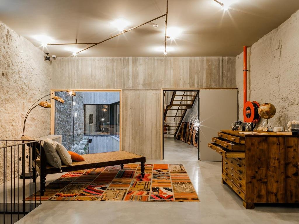 A Guide to the Best Day Tours From Porto An industrial-chic room interior at Armazém Housing in Porto, featuring a polished concrete floor, exposed stone walls, a low wooden daybed on a colorful geometric patterned rug, and a large vintage wooden chest of drawers under warm ceiling track lighting.