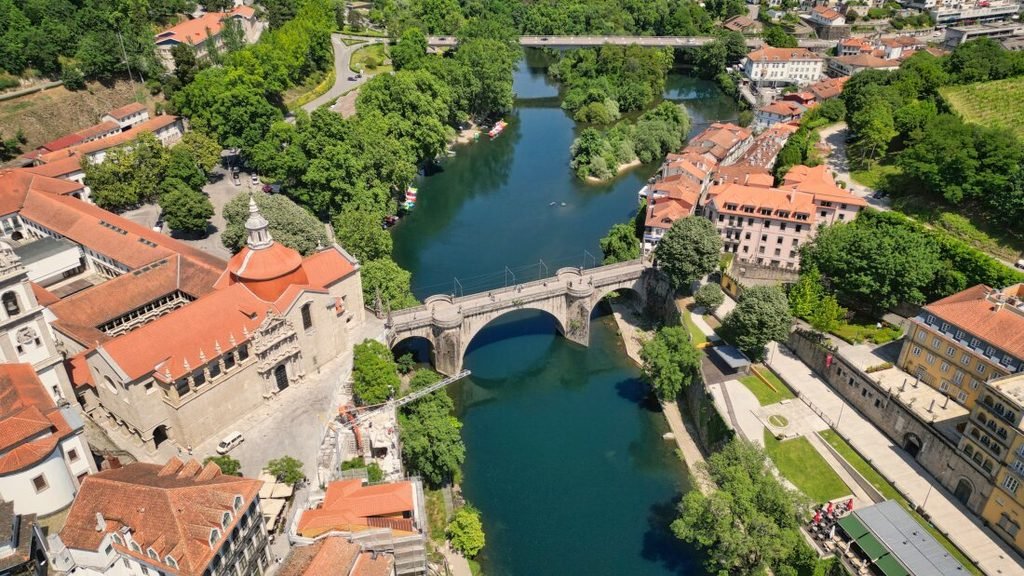 Best Towns in Northern Portugal: A Photographer’s Map A beautiful high-angle aerial view of Amarante on a bright sunny day, featuring the historic stone São Gonçalo Bridge arching over the clear teal waters of the Tâmega River next to the grand red-domed São Gonçalo Church, all surrounded by lush green trees and terracotta-roofed buildings.