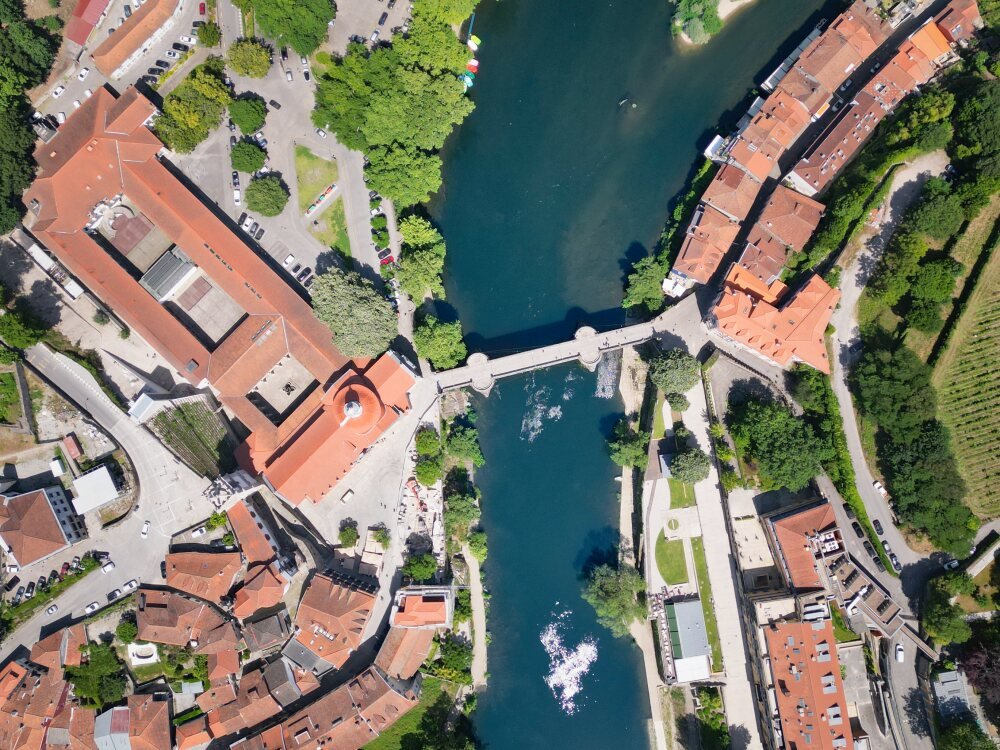 A dramatic top-down drone aerial photograph of Amarante, showcasing the historic stone São Gonçalo Bridge spanning across the deep blue waters of the Tâmega River, with the large orange-tiled roof complex of the São Gonçalo Church on the left bank and narrow terracotta-roofed streets framing the right bank