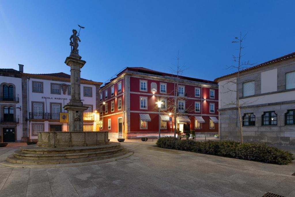 Best Towns in Northern Portugal: A Photographer’s Map A beautiful evening wide shot of a public stone square in Viana do Castelo, featuring an ancient granite fountain pillar statue in the foreground and the illuminated red historic facade of the AP Dona Aninhas hotel building under a deep blue twilight sky.