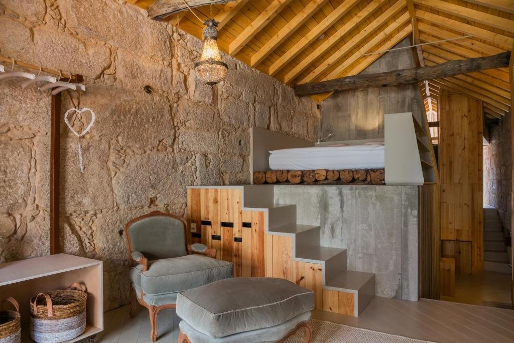 Best Towns in Northern Portugal: A Photographer’s Map A unique hotel loft room at Armazém Luxury Housing featuring a high vaulted wooden ceiling, exposed stone walls, a grey velvet armchair with an ottoman, and a raised bed platform accessible by a modern wooden staircase.