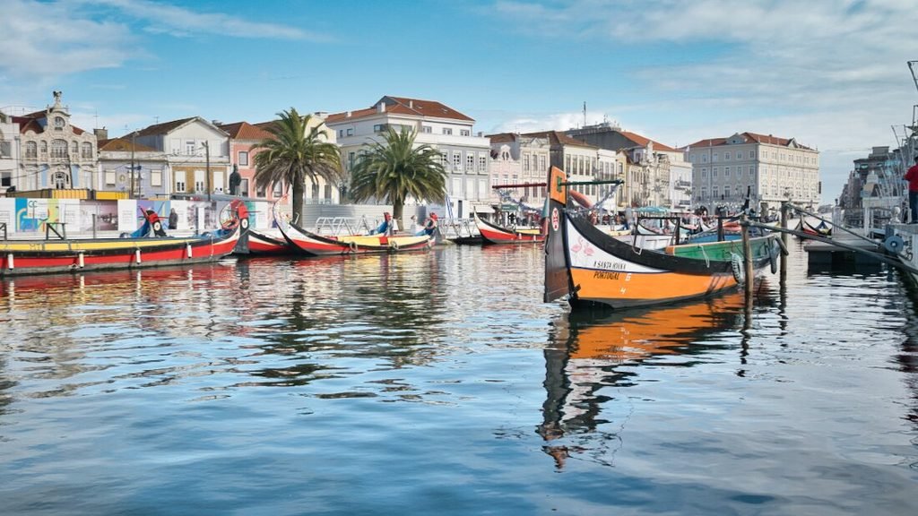 A Guide to the Best Day Tours From Porto A vibrant orange moliceiro boat floating along a saltwater canal in Aveiro on a bright sunny day under completely clear blue skies.