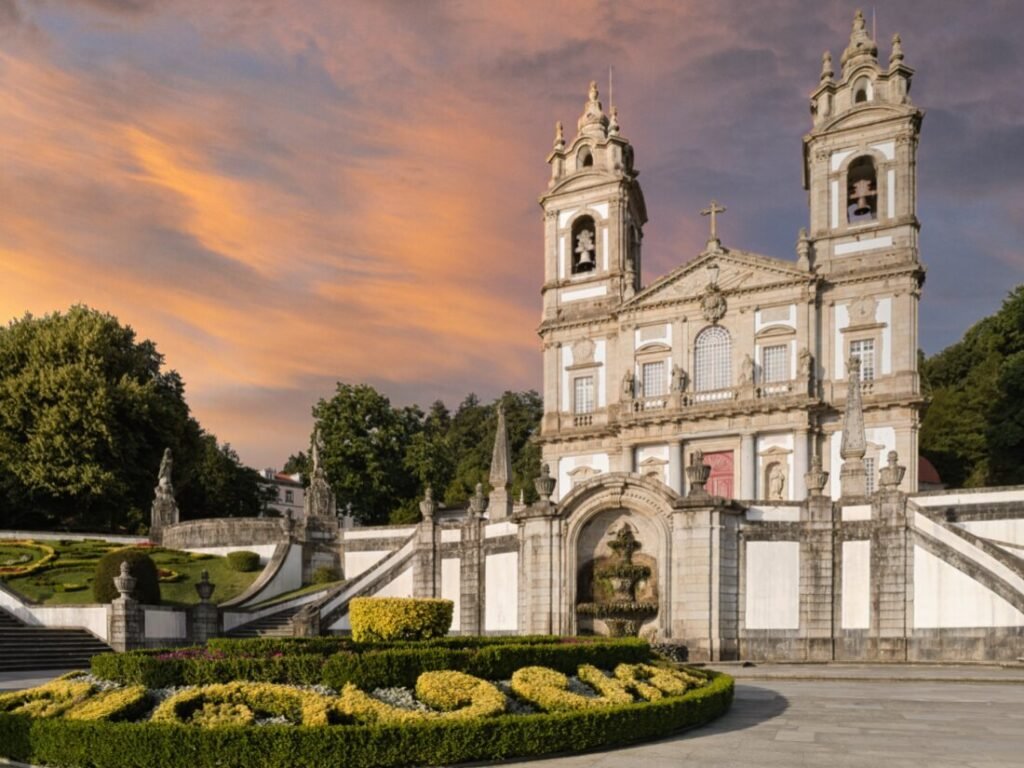 The grand church facade of the Bom Jesus do Monte sanctuary in Braga at twilight, featuring a deep red evening sky in the background and manicured green gardens in the foreground