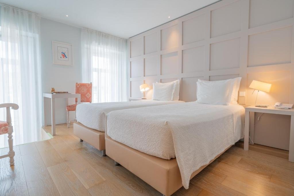 Best Towns in Northern Portugal: A Photographer’s Map A bright and minimalist boutique hotel guest room at the Burgus Tribute & Design Hotel, featuring twin beds with crisp white linens, light oak wood floors, a grid-patterned accent wall, and soft white curtains filtering natural daylight.