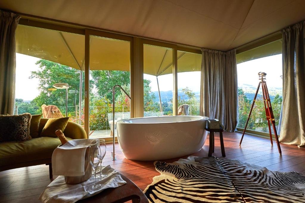 Best Towns in Northern Portugal: A Photographer’s Map The luxurious interior of a safari-style tent suite at Carmo's Boutique Hotel, featuring a freestanding white bathtub in the center of hardwood floors, a zebra print rug, a velvet sofa with champagne in an ice bucket in the foreground, and floor-to-ceiling glass windows opening onto a private terrace with views of lush green rolling hills