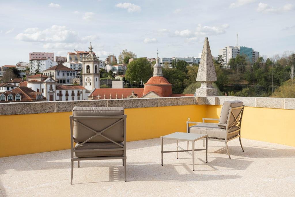 Best Towns in Northern Portugal: A Photographer’s Map A spacious rooftop terrace at Casa da Calçada in Amarante, featuring two modern grey lounge chairs and a small table on a stone tiled floor, set against a yellow wall with panoramic views of the historic town, church spires, and terracotta roofs