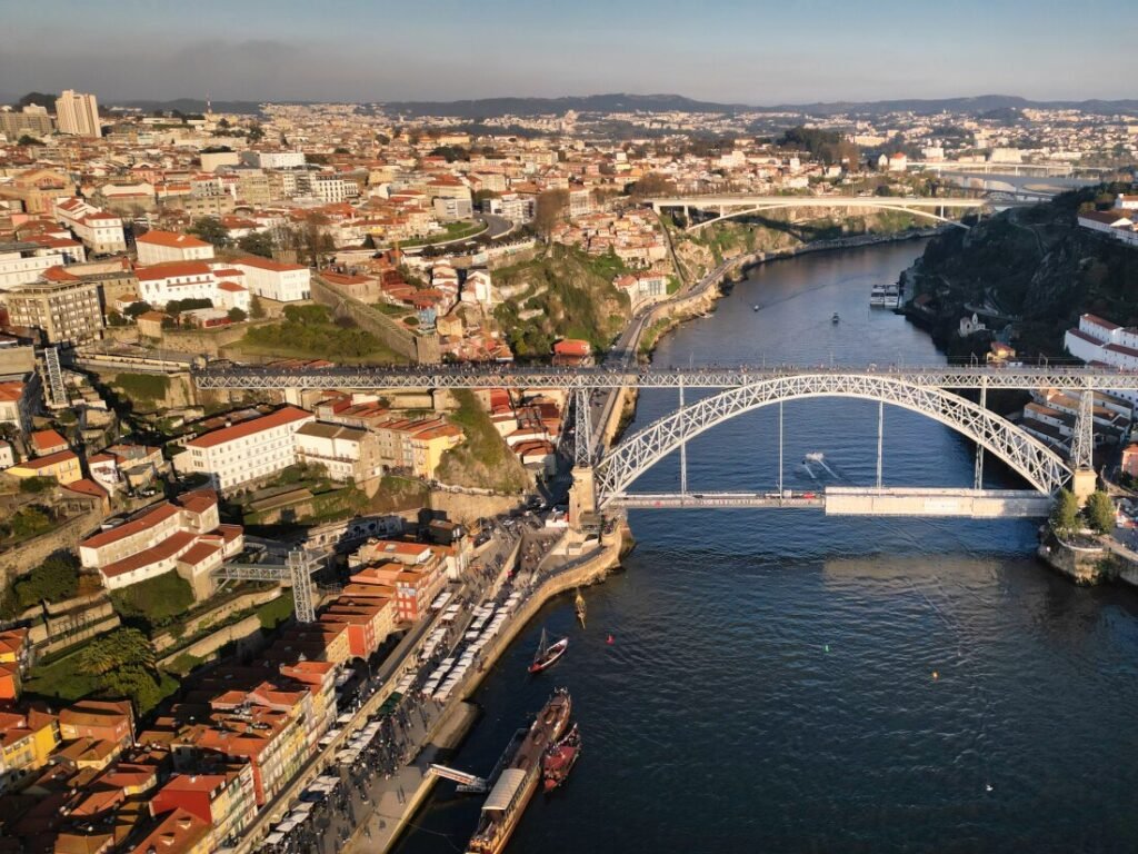 A spectacular aerial drone view looks down on the historic Dom Luis I Bridge spanning the Douro River in Porto, with a traditional wooden rabelo boat gliding along the water on the left.