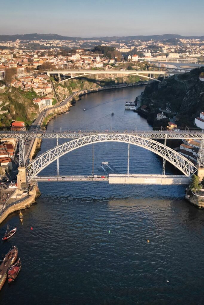 A spectacular aerial drone view looks down on the historic Dom Luis I Bridge spanning the Douro River in Porto, with a traditional wooden rabelo boat gliding along the water on the left.