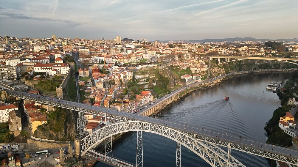 Best Towns in Northern Portugal: A Photographer’s Map An expansive, high-angle aerial view of the double-deck metal arch Dom Luís I Bridge over the Douro River in Porto, showcasing a boat sailing up the river and the sprawling cityscape climbing the hills under soft late-afternoon skies.