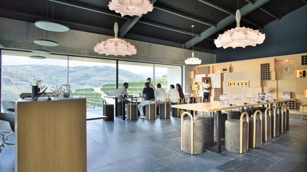 A Guide to the Best Day Tours From Porto An interior view of a modern, renovated wine tasting room featuring a sleek layout, with expansive windows displaying rolling green vineyards and layered mountains in the background