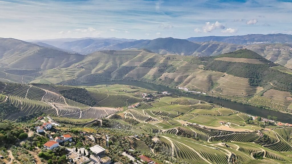 Solo Female Travel in Porto: 3 Day Slow-Travel Itinerary A spectacular aerial drone perspective looks directly down on the shimmering blue curves of the Douro River winding past dramatic emerald green terraced vineyards under a clear blue sky.
