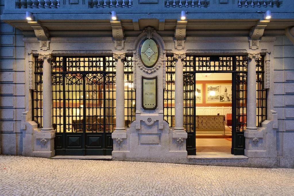 Best Towns in Northern Portugal: A Photographer’s Map The grand stone facade and entrance of the EMAJ Guimarães Boutique Hotel, featuring historic neoclassical architecture, intricate black wrought-iron gates, stone columns, and warm exterior downlighting on traditional Portuguese pavement.