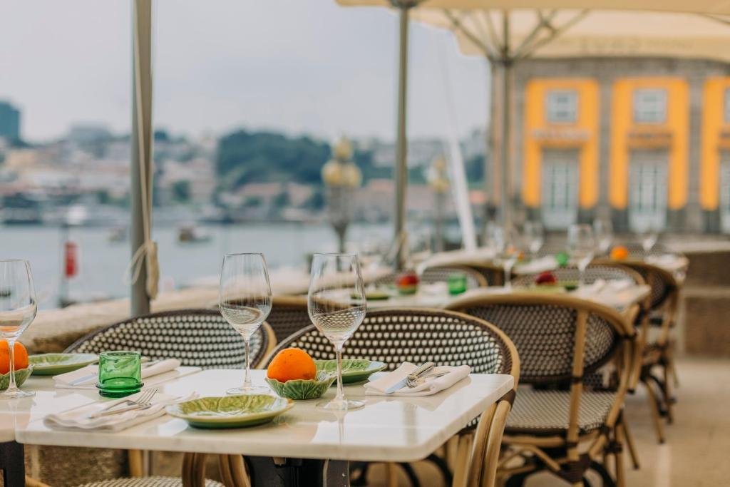 Best Towns in Northern Portugal: A Photographer’s Map An elegant outdoor restaurant dining terrace overlooking the river, featuring set tables with wine glasses, woven chairs under a large umbrella, and a historic yellow building in the blurred background