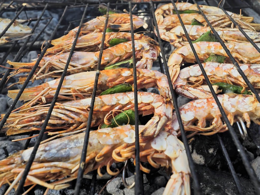 A close-up photograph of large prawns sizzling and charring beautifully on a metal grill over hot charcoal ashes.