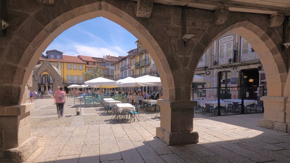 Best Towns in Northern Portugal: A Photographer’s Map Uma vista emoldurada por arcos de pedra medievais a partir do alpendre do antigo palácio municipal na Praça de Santiago, em Guimarães, revelando esplanadas cobertas por chapéus de sol brancos, edifícios históricos de fachadas coloridas e o padrão tradicional do pavimento de granito sob um céu azul