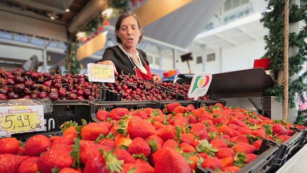 Solo Female Travel in Porto: 3 Day Slow-Travel Itinerary A friendly local market vendor smiles behind a vibrant, overflowing stall completely packed with fresh, bright red strawberries inside the historic Mercado do Bolhao in Porto.