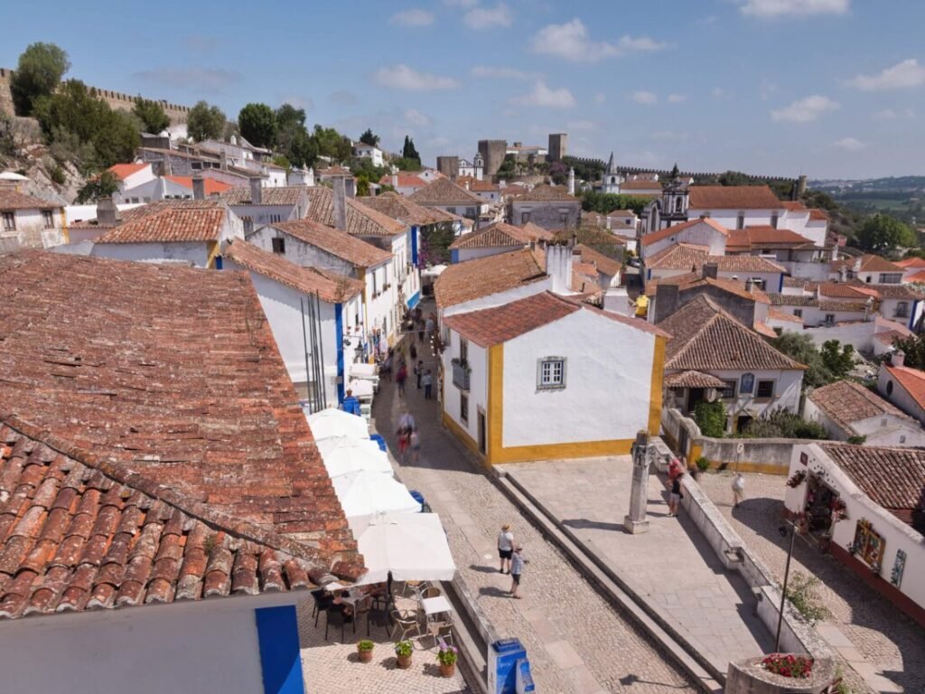 A scenic view looking down from the historic stone walls of Óbidos castle over a narrow cobblestone street lined with traditional whitewashed houses.