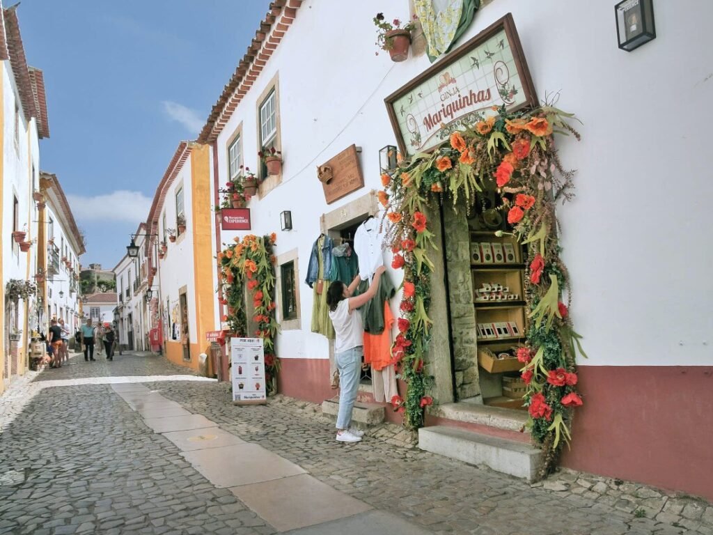 A local shop owner setting up a display of handmade crafts and artisanal goods outside a traditional white-walled storefront on a cobblestone street in Óbidos.
