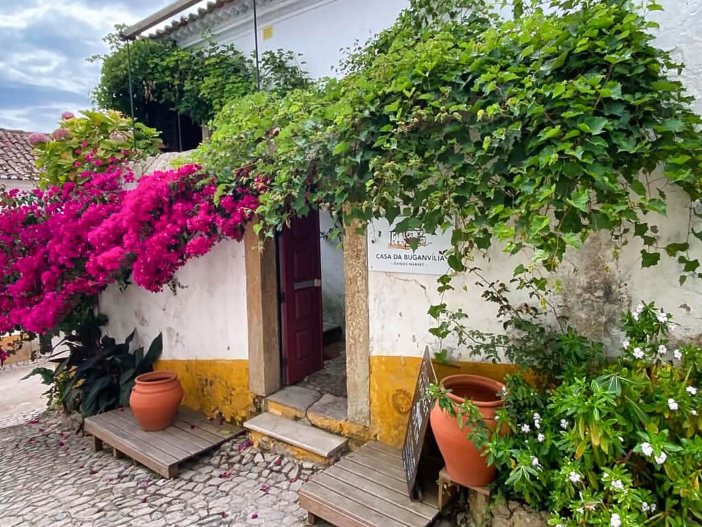 A stunning bright pink bougainvillea plant climbing the facade of a traditional whitewashed house with vibrant yellow borders along a quiet cobblestone street in Óbidos on a sunny day.