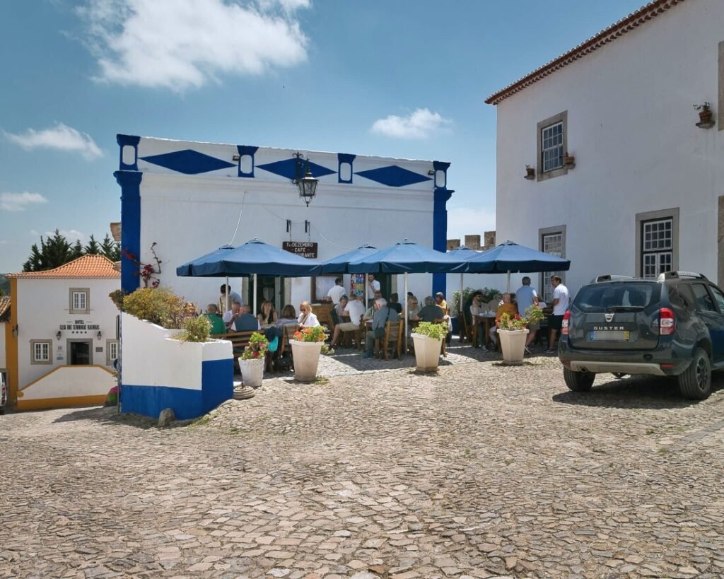 An inviting outdoor restaurant terrace on an Óbidos cobblestone street, featuring guests dining under vibrant blue parasols alongside traditional whitewashed buildings on a sunny day.