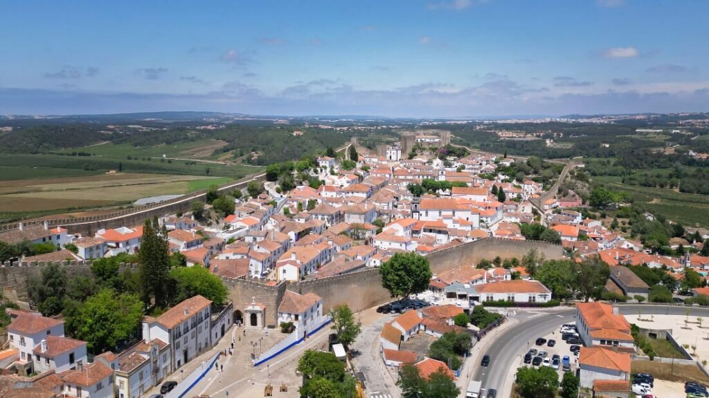 A stunning aerial drone view looking down on the complete medieval castle walls and historic whitewashed village of Óbidos under a clear blue sky.