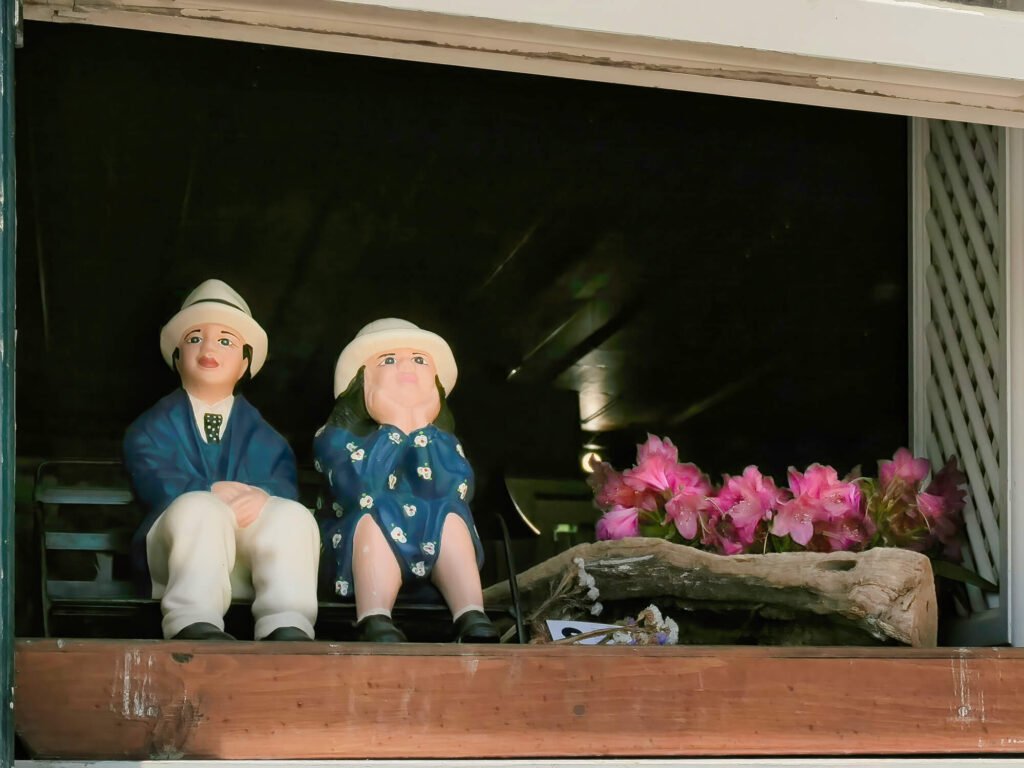 Two small, traditional ceramic couple figurines sitting side-by-side on an open window sill overlooking a scenic street inside the medieval village of Óbidos.