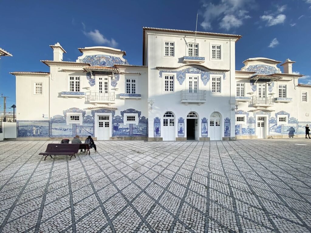 The Poetry of the Silver Coast, Portugal The historic white facade of the old Aveiro railway station, lavishly adorned with traditional Portuguese blue and white azulejo tile panels depicting regional motifs under a clear sky.