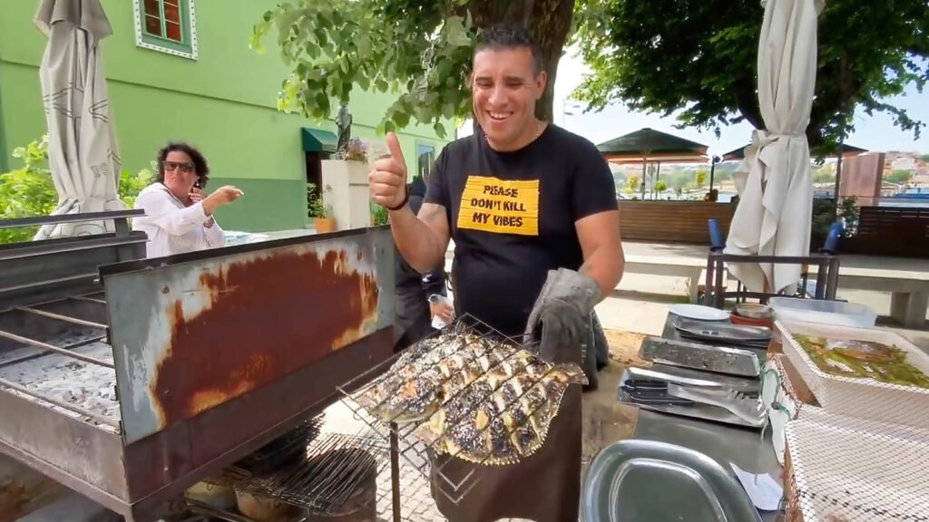 The owner Pedro standing proudly at Casa Moreira in the fishing village of Afurada, holding up a freshly charred whole fish directly over a smoking outdoor charcoal grill.