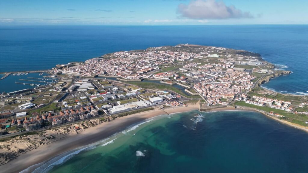 The Poetry of the Silver Coast, Portugal A stunning, wide aerial drone photograph of the entire Peniche peninsula jutting out into the deep blue Atlantic Ocean on a bright, clear sunny day.