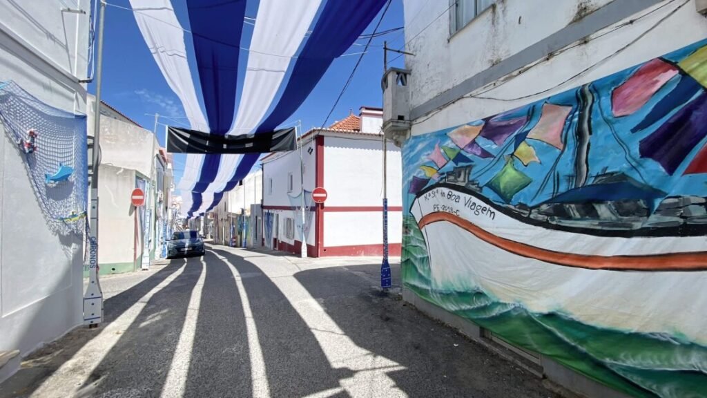 The Poetry of the Silver Coast, Portugal A sun-drenched, narrow cobblestone street in Peniche lined with traditional whitewashed houses adorned with expressive graffiti street art, shaded by geometric blue and white sails stretched overhead against a bright summer sky