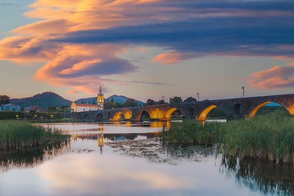 Best Towns in Northern Portugal: A Photographer’s Map A stunning low-angle twilight landscape photograph of Ponte de Lima, featuring the historic stone Roman bridge with its ancient arches illuminated in a warm golden glow, perfectly reflected in the calm river waters beneath a dramatic sky filled with vibrant pink and orange clouds