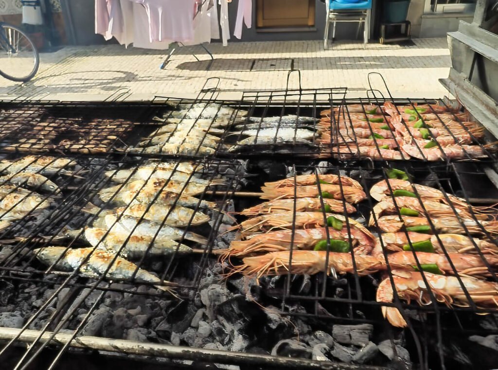Fresh Atlantic sardines and large prawns sizzling over a traditional open-air charcoal grill box with light smoke rising from the hot embers.