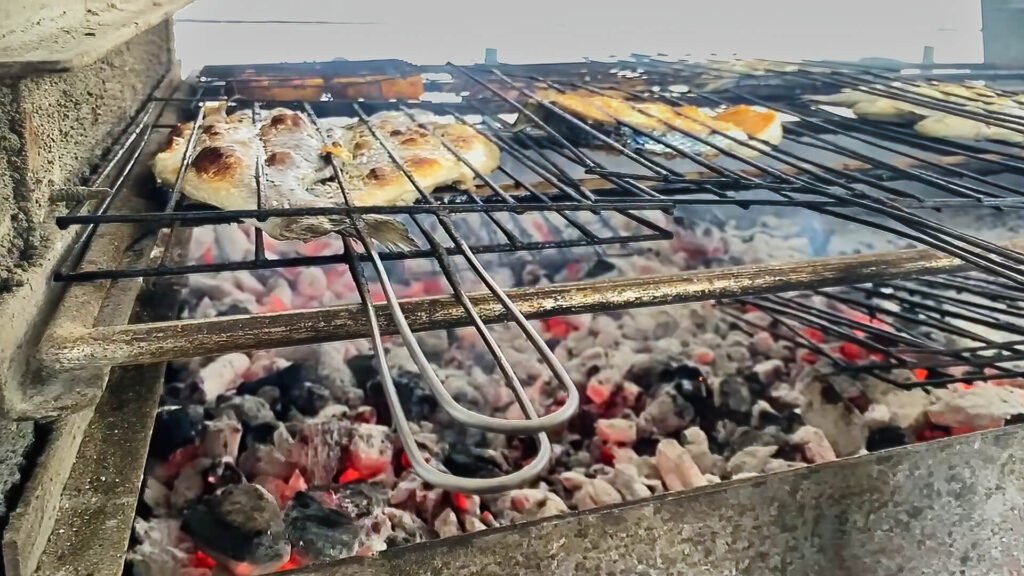 A macro close-up of fresh whole fish crisping beautifully directly over hot, glowing charcoal embers on an open-air barbecue grill.