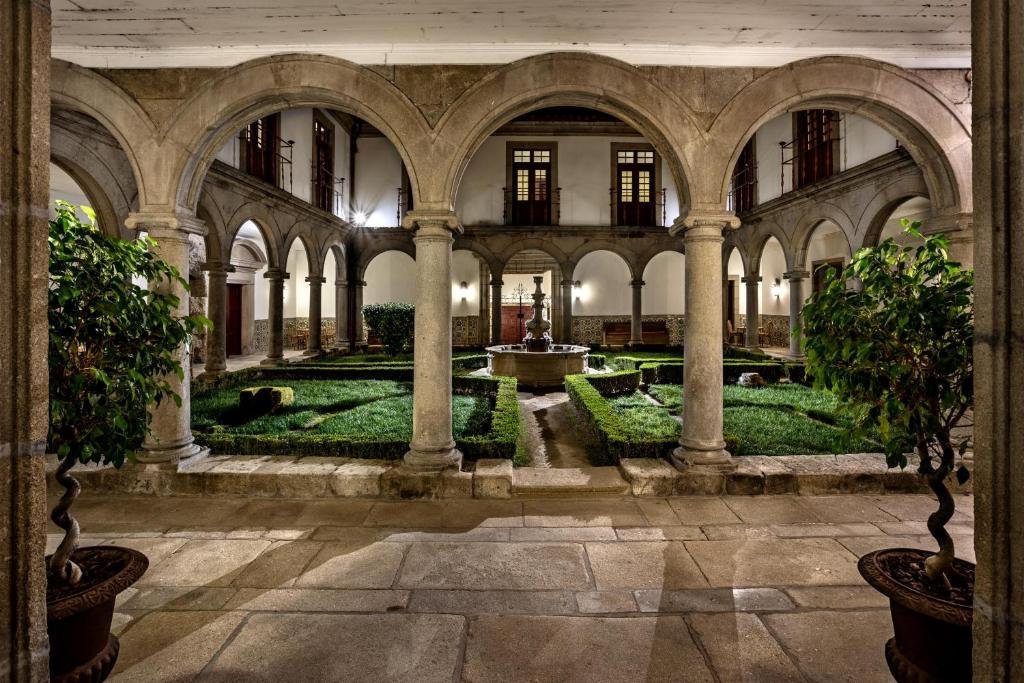 Best Towns in Northern Portugal: A Photographer’s Map A historic stone courtyard cloister arcade at the Pousada Mosteiro de Guimarães hotel, featuring massive granite arches supported by columns, a central fountain surrounded by manicured hedges, and warm evening lighting.