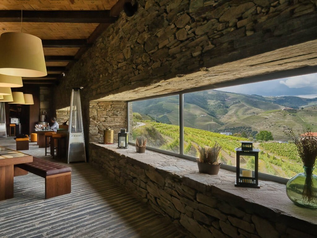 Douro Valley Harvest, Portugal: Best Stays & Dining The modern wine tasting room inside Quinta do Val Moreira, featuring a rustic stone accent wall framing a large glass window that looks out over the cascading terraced vineyards of the Douro Valley.