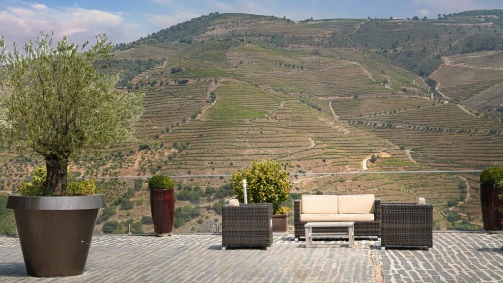 Douro Valley Harvest, Portugal: Best Stays & Dining An open-air terrace at Quinta do Val Moreira featuring a comfortable lounge sofa and a potted olive tree, overlooking the expansive mountain ridges and terraced hills of the Douro Valley.